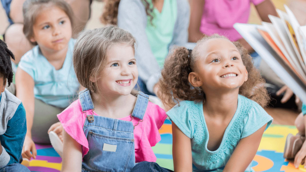 Two girls sitting on a carpet and smiling.